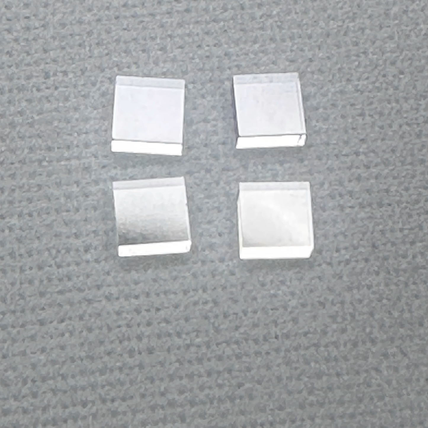 Four square-shaped, silver-colored diamond single crystal wafers/substrates arranged on a textured grey surface.